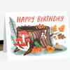 A greeting card with the text "HAPPY BIRTHDAY" above an illustration of a woodland tree stump, red mushrooms, autumn leaves, and a small green frog.