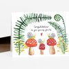 A folded greeting card with a watercolor illustration of three red mushrooms with snails, green fern leaves, small yellow flowers, and the text "Congratulations on your growing family" on the front.