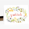 A folded greeting card with a watercolor wreath of leaves, berries, acorns, and mushrooms around the word “gratitude” in coral script.