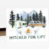 A greeting card with a watercolor camping scene showing a trailer, station wagon, pine trees, mountains, and two camp chairs around a fire, with the text "HITCHED FOR LIFE".