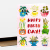 A folded greeting card with a pink front featuring illustrated potted plants, a green frog, and the red text "HAPPY BIRTH DAY!".