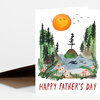 A folded Father’s Day greeting card with an illustrated coastal scene of pine-covered cliffs, whales in the water, an orange sun, and the text "HAPPY FATHER'S DAY" on the front.