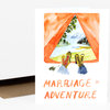 A folded greeting card shows a watercolor illustration of a tent opening to a mountain landscape with boots and the text "MARRIAGE = ADVENTURE" on the front.