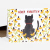 A folded greeting card shows the red words "NEVER FORGOTTEN" above a black cat silhouette with a red collar, surrounded by yellow flowers.