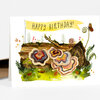 A folded greeting card with a watercolor-style illustration of a log covered in mushrooms and plants, a butterfly at the upper right, and the text “HAPPY BIRTHDAY!” on a yellow banner.
