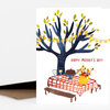 A folded greeting card shows an illustrated picnic table under a dark blue tree with yellow leaves, string lights, flowers, wine glasses, cake, and the text "HAPPY MOTHER'S DAY!".