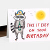 A folded birthday card featuring an illustrated raccoon holding a bottle, a yellow sun, small birds, and the text "TAKE IT EASY ON YOUR BIRTHDAY" in red.