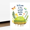 A folded greeting card with the text "Welcome to the world little one" in blue script, surrounded by watercolor butterflies, fern-like plants, mushrooms, and a green sprouting mound with roots.