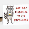 A folded greeting card shows a watercolor raccoon holding a takeaway coffee cup next to the red text "YOU ARE ESSENTIAL TO MY HAPPINESS".