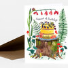 A folded greeting card featuring a watercolor illustration of a yellow layer cake topped with blackberries and candles, surrounded by plants, mushrooms, and a snail, with the text "Happiest of Birthdays" on the front.