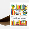 A folded greeting card with a watercolor illustration of bookshelves, potted plants, and paintbrush mugs, and the handwritten message "Wishing you books and more books on your birthday".