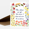 A folded greeting card with a watercolor floral border and the hand-lettered text "The older we get, the more I cherish our friendship" on the front.