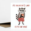 A white Valentine’s Day greeting card with a raccoon illustration holding a remote and a drink, with the text "IT'S VALENTINE'S DAY" at the top and "LET'S DO THIS" at the bottom.