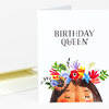 A folded greeting card with the text "BIRTHDAY QUEEN" and a colorful illustrated face wearing a flower crown on the front.