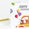 A folded birthday notecard with the text "HAPPY BIRTHDAY" and watercolor illustrations of balloons and two dogs in a vehicle.