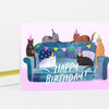 A folded birthday card with a pink front showing six cats wearing party hats on a blue couch, colorful bunting, and the text "HAPPY BIRTHDAY!".