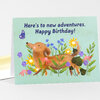 A folded birthday card with the text "Here’s to new adventures. Happy Birthday!" and an illustration of a brown dog standing among colorful flowers and a butterfly.