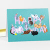 A folded birthday card with a teal front printed with pastel purple "HAPPY BIRTHDAY" text and illustrated cats wearing party hats.