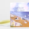 A folded birthday greeting card with the text "BIRTHDAY GREETINGS" and an illustrated seaside scene featuring several seagulls, flowers, and a sandy beach.