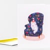A folded greeting card featuring an illustration of a white cat wearing a party hat, sitting on a dark blue floral armchair with a pink oval rug beneath it.