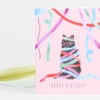 A pink birthday greeting card shows a black cat wrapped in colorful streamers with the words "HAPPY BIRTHDAY" at the bottom.
