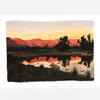 A postcard featuring a painted landscape of red-orange mountains, dark trees, and their reflection in still water at sunset.