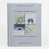 A hardcover book with a light gray cover titled “YOU ARE A SACRED PLACE,” with the subtitle “VISUAL POEMS FOR LIVING IN CLIMATE CRISIS” and the author name “MADELEINE JUBILEE SAITO,” plus a four-panel illustration of leaves, raised hands, a reclining figure, and berries.