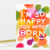 A folded greeting card with colorful confetti-like blobs and the white hand-lettered text "I'M SO HAPPY YOU WERE BORN" on the front.