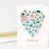 A folded greeting card with a white front showing a heart shape made of blue and green vines and orange flowers, with the words "thank you" printed at the bottom.