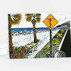 A folded greeting card with a colorful illustrated scene of a beach road, palm trees, flowers, a bicycle, and a yellow diamond road sign with a surfer silhouette.