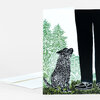 A folded greeting card with a linocut-style illustration of a black dog looking up at a person’s legs in black pants and shoes among green trees and grass.