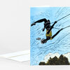 A folded greeting card shows an illustrated person swinging upside down by ropes against a blue sky above trees.