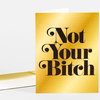 A folded greeting card with a metallic gold front printed in large black serif text reading "Not Your Bitch," with a white envelope partially visible behind it.