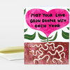 A folded greeting card with a large pink heart-shaped flower and the text "May Your Love Grow Deeper With Each Year," with green leaves and illustrated roots below.