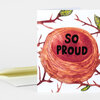 A folded greeting card with a illustrated bird nest and branches design and the words "SO PROUD" in bold black letters on the front.