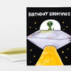 A birthday greeting card with the text "BIRTHDAY GREETINGS" and an illustration of a green alien in a silver flying saucer beaming up a yellow roadway against a black starry design.