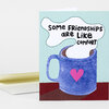 A folded greeting card shows a blue mug with a pink heart and the text "SOME FRIENDSHIPS ARE LIKE COMFORT" in bold black letters.