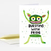 A folded greeting card with a whimsical green creature illustration and the text "BURSTING WITH PRIDE" on the front.
