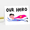 A folded greeting card with the words "OUR HERO" and an illustration of a flying superhero in a blue outfit with a pink cape and yellow star.