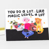 A folded greeting card with the text "YOU DO A LOT... LIKE MAGIC LEVEL A LOT" above a colorful illustration of a person sitting with a laptop, mug, basket of plants, and a yellow dog.