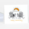 A folded anniversary greeting card with a line drawing of two people sitting in Adirondack chairs facing a sun over mountains and the words "happy anniversary" in orange lettering.