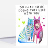 A folded greeting card with the text "SO GLAD TO BE DOING THIS LIFE WITH YOU" above two colorful illustrated animals and small heart shapes.