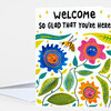 A greeting card with the text "WELCOME SO GLAD THAT YOU'RE HERE" above colorful illustrated flowers, leaves, and a bee.