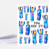 A folded greeting card with illustrated graduates in blue caps and gowns surrounding the text "YOU DID IT!" on the front.