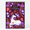 A spiral-bound wall calendar cover with the text "papio press 2027 CALENDAR" and "PAPIOPRESS.CO.UK," featuring a white unicorn surrounded by purple flowers and pink leaves.