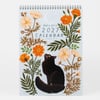 A spiral-bound 2027 calendar with botanical flower illustrations surrounding a black cat and the text "papio press 2027 CALENDAR" on the cover.