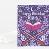 A folded greeting card with the words "Happy Birthday Witch" on the front, illustrated with a crescent moon, stars, an open spellbook, crossed broomsticks, mushrooms, and leafy floral decorations in purple, pink, and lavender.