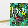 A greeting card with a black background covered in green and teal jungle leaves, large white text reading "SENDING HUGS!", and an illustration of a person hugging a bear.