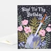 A folded birthday card with a black front reading "Slay! Tis Thy Birthday" in white gothic lettering above floral artwork, a sword, and a stone pedestal.