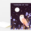 A folded greeting card with a dark cover showing the text "THINKING OF YOU," a white owl, a moon, stars, and purple flowers, with a blank white envelope behind it.